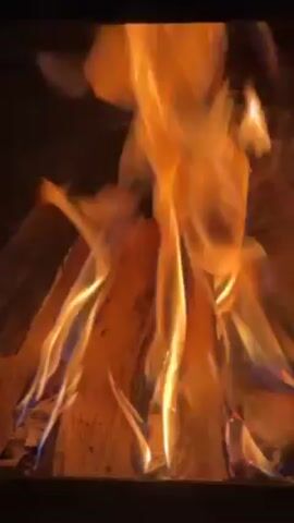 The video shows a close-up view of a fire burning inside a fireplace or wood-burning stove. Logs are arranged in the firebox, with flames rising and dancing around them. The flames are predominantly orange and yellow, with some hints of blue. The background is dark and indistinct. The camera is steady, focusing on the movement of the fire.