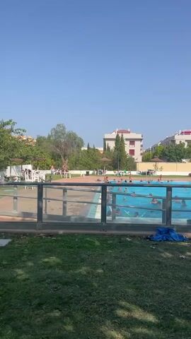 A large, outdoor swimming pool is filled with clear blue water. Numerous people are visible in the pool, some swimming and others interacting with each other. A blue water slide with white accents is situated at one end of the pool, with a few individuals at its entrance. Surrounding the pool is a paved area with lounge chairs, some occupied by people relaxing. Beyond the pool, lush green trees and grass are present, interspersed with buildings in the background. A clear blue sky dominates the upper portion of the frame. The video appears to be filmed from a slightly elevated position, looking down at the pool area.
