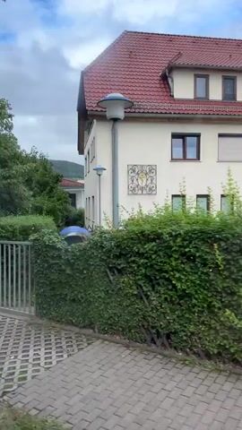 The video begins by showing a sign with red lettering that reads "Sa. 10 Uhr". The camera then pans to reveal a light-colored building with a red tiled roof. Adjacent to this building is a blue building. In front of the blue building, several cars are parked in a lot paved with interlocking bricks. A black electric vehicle charging station is also visible. The blue building has several windows and a sign that says "A Apotheke von Hirschhausen". The video continues to show the blue building and the parking lot. The sky is overcast.