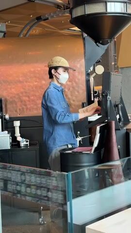 A person wearing a denim shirt, a light-colored baseball cap, and a white face mask stands in front of a large coffee roasting machine. The person is wearing grey pants and is looking at papers they are holding. They are operating the machine with their right hand. The machine is black and copper and has a large hopper at the top. There is a glass partition in the foreground with shelves of products behind it.