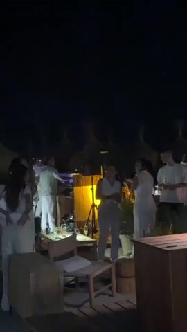 A group of people dressed in white are gathered outdoors at night. They appear to be at a party or event, with some individuals dancing and interacting. There is a DJ booth with speakers and lighting, suggesting music is playing. In the background, there are structures with thatched roofs, possibly part of a resort or beach establishment. The overall atmosphere seems festive.