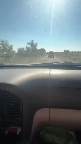 The video is taken from the interior of a vehicle, looking out through the windshield. The camera perspective suggests the viewer is in the passenger seat or the driver's seat. The dashboard of the vehicle is visible in the foreground. Ahead, a dirt road stretches into the distance, characterized by numerous potholes and uneven surfaces. The road is lined with sparse vegetation and dry, scrubby bushes. In the distance, another dark-colored SUV is traveling in the same direction. The sky is clear and bright blue, with the sun high overhead, causing a lens flare effect that streaks across the upper part of the frame. The overall impression is of a journey on a rough, rural road under a sunny sky.