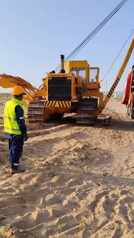 A man wearing a yellow hard hat and a high-visibility vest stands facing a large yellow construction vehicle. The vehicle is a tracked machine, possibly a dozer or a pipelayer, with a blade at the front and a boom extending upwards and to the right. The boom is supporting a large pipe, which is being lifted from a flatbed truck. The truck is red and loaded with several large, dark pipes. The scene appears to be an outdoor construction or industrial site, with sandy terrain and a clear blue sky. The man is observing the operation of the heavy machinery.