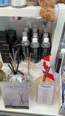 The video pans across shelves filled with candles and related products in a retail setting. On the top shelf, various colors of long taper candles are displayed in glass jars. Below this, on the second shelf, are several bottles of what appear to be essential oils or fragrance diffusers, arranged neatly in rows. Some boxes labeled "Everlasting Candle Co." are stacked on the left. Also visible are reed diffusers with various colored reeds. On the lower shelves and counter space, more candles are displayed, including pillar candles, teardrop-shaped candles, and tealights in packaging. There are also decorative candle holders, some made of glass and others metallic gold. A small Canadian flag is positioned near some of the diffuser bottles. The environment appears to be a well-lit store with white shelving. No people are clearly visible in the video, though a faint reflection of a person's head and shoulders can be seen in one of the glass jars on the top shelf. The camera is handheld and moves smoothly, providing a comprehensive view of the merchandise.