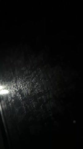 The video shows a dark scene, likely viewed through a rain-streaked window. A bright light source is visible at times, creating reflections and glare on the water droplets. Occasional blurry movements suggest the camera may be in a moving vehicle. There are no discernible people or objects beyond the window and the light. The overall environment is dark and wet.