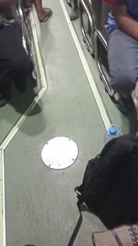 The video provides a view from inside a vehicle, likely a bus or train, looking down an aisle between rows of seats. Several passengers are visible, some seated and others standing further down the aisle. The floor is a light green color with yellow and white striped markings delineating the aisle. In the foreground, a round metal plate is set into the floor. The camera pans slowly, showing a mix of seated passengers in casual clothing, some looking at their phones, others appearing to rest. The lighting suggests an indoor environment, possibly during the day, with a muted color palette. The overall impression is one of ordinary travel.