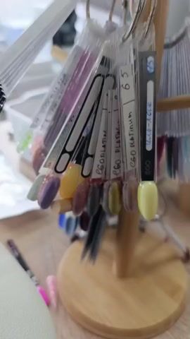 This video shows a close-up of a nail polish display stand. The stand is made of wood and holds multiple sets of nail polish swatches on rings. The swatches are oval-shaped and are displayed in a fan-like arrangement. Each swatch has a label indicating the color and brand. The background is blurred, but appears to be an indoor setting, possibly a nail salon, with other displays and equipment visible. The lighting is bright and natural, highlighting the colors of the nail polishes. The video is shot from a slightly elevated angle, looking down at the display.