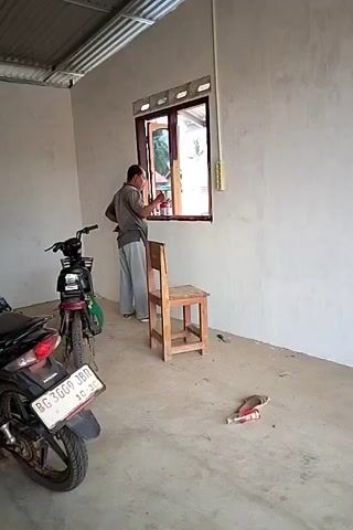 A man in a grey t-shirt and light grey pants is standing by a window in what appears to be an unfinished room. He is holding a red object, possibly a paint can, and a brush in his hands and appears to be painting the window frame. To his left, there are two motorcycles parked. One is a black motorcycle with a license plate that reads "BG 3009 JBO". To his right, there is a wooden chair. On the floor near the chair, there are two bottles, one red and one clear, and a pair of sandals. The room has bare concrete floors and white walls. The ceiling is corrugated metal. The window is open and shows greenery outside.