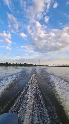A first-person perspective video shows the wake of a boat traveling down a wide river. The water is dark with frothy white streaks of the wake. The sky is a vibrant blue with scattered, puffy white clouds. On the left bank of the river, there is a distant line of green trees. In the distance on the right, there is a tall, thin tower, possibly a radio tower, and a bridge can be seen. The lower left corner of the frame shows a portion of the back of the boat. The video is taken from the perspective of someone sitting in the boat.
