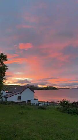 A slow-motion video shows a pink and orange sunset over a body of water. In the foreground, a white house with a red roof is visible on a green hillside. Trees are on the left and right sides of the frame. The sky is filled with clouds, and the sun is setting behind mountains in the distance. The water is calm and reflects the colors of the sunset.