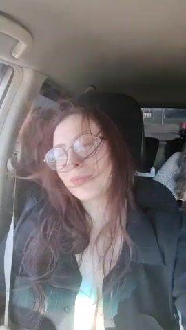 The video shows a young woman in the front passenger seat of a car. She has long, reddish-brown hair, round glasses, and is wearing a dark jacket over a light-colored top. Her hair is blowing around her face. In the background, another woman is visible in the back seat, looking at her phone. The interior of the car is dimly lit, and the view outside the window suggests it is daytime. The woman in the front seat occasionally smiles and moves her head, causing her hair to become more disheveled. There are glimpses of the car's dashboard and seatbelts. The video is shot from a selfie perspective, indicating the woman is filming herself.