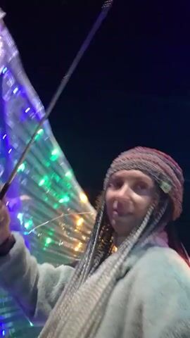 This is a short video clip featuring a person dressed in a light blue hoodie and a grey knit hat. The person has long, braided hair. They are holding an umbrella with a long, thin metal handle, and the umbrella is decorated with colorful LED lights that flash in green, blue, and purple. The background is dark, suggesting it is nighttime. The scene appears to be outdoors, possibly at an event or festival, given the decorative lights. The person is looking towards the camera with a slight smile. The text "00:00" to "00:04" appears overlaid on the footage, indicating timestamps. No other text is visible. The overall mood is festive and cheerful.