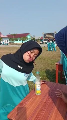 A young woman wearing a black hijab and a light blue and teal shirt attempts to pick up marbles from a bottle using a straw. She is positioned over a wooden table, with the bottle in front of her. Her cheeks are puffed out as she tries to create suction with the straw. In the background, other young women in similar teal outfits are visible, some standing and watching, others participating in activities on a grassy field. A building with a sign is visible in the distance. The sky is clear and blue.