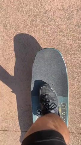 The video shows a person riding a skateboard down a sidewalk. The person's legs and the skateboard are visible in the first-person point-of-view shot. The skateboard has the words "EL MUNDO QUE VIN" printed on its side. The sidewalk is made of concrete with some red sections. The video then pans up to show the sidewalk stretching into the distance, lined with trees on one side. A street with parked cars is visible on the left, and a concrete structure is on the right. The day appears sunny and clear.
