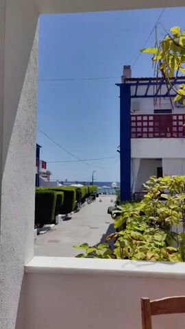 The video shows a view from an opening, looking out onto a pathway leading towards the sea. On the left, there is a white wall and a neatly trimmed green hedge. On the right, there are buildings with white, red, and blue architectural elements, and green foliage. The pathway is paved and lined with hedges. In the distance, the sea is visible under a clear blue sky. There are no people or specific actions clearly depicted in the frame.