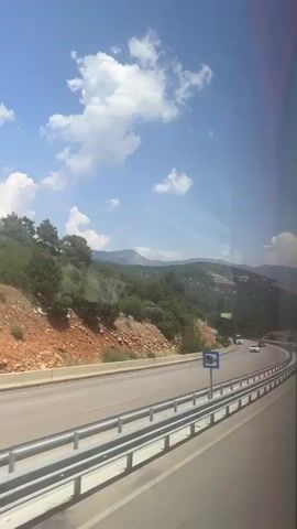 This is a shot from a moving vehicle, looking out a window at a highway winding through a mountainous, tree-covered landscape under a bright blue sky with fluffy white clouds. A metal guardrail runs along the edge of the highway. Several white cars and a white truck with a black tarp-covered trailer are visible driving on the road. The terrain consists of steep, rocky slopes covered in green trees and vegetation. The sun is shining, creating bright highlights and shadows.