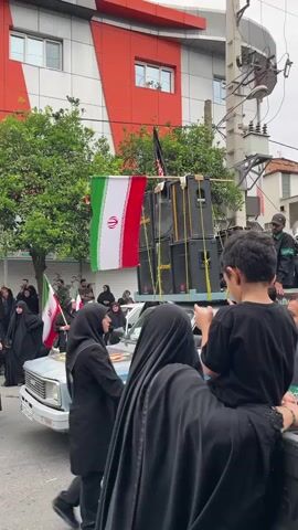 A parade is taking place in an urban setting in Babol, Iran. A light blue pickup truck is the central object, adorned with an Iranian flag and a large sound system comprising multiple speakers mounted on top. The truck is moving forward, likely participating in the procession. Several people are visible in the scene, most of whom are wearing black clothing, including hijabs and chadors, suggesting a somber or religious event. A young boy in a black t-shirt is seated in the back of the truck, looking towards the right. An adult, also in black clothing and a hijab, is seated next to the boy. In the background, more people are walking, many holding Iranian flags. The buildings in the background appear to be commercial or residential, with one building having a striking orange and white facade. Trees line the street, and utility poles with wires are present. The sky is overcast, suggesting a cloudy day. The overall atmosphere appears to be one of organized gathering and possibly religious observance. No specific text is clearly legible on the visible objects or clothing.