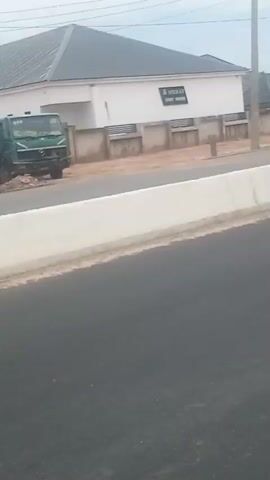 A green truck is parked in front of a white building with a dark roof. A person is standing on the side of the road. A yellow auto-rickshaw drives past. A silver car is driving on the road. The scene appears to be a street in a town or city with buildings and a road.