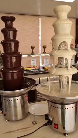The video shows two chocolate fountains side-by-side. The fountain on the left is filled with dark chocolate, and the fountain on the right is filled with white chocolate. Both fountains are cascading chocolate down multiple tiers. In the background, there is a buffet table with fruit and other food items. The scene appears to be indoors, possibly in a restaurant or banquet hall, with blinds covering a window behind the buffet. There are no people actively interacting with the chocolate fountains in this clip.