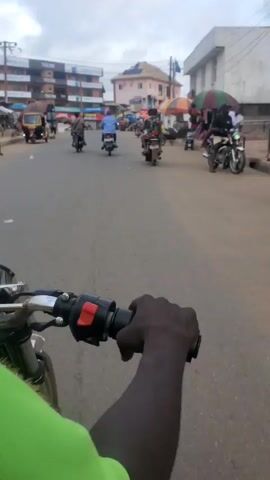 The video shows a first-person perspective of someone riding a motorcycle on a street. Several other motorcycles are driving in the same direction, further down the road. There are a few small commercial buildings on either side of the street with various signage, including several for “TECNO.” There are vendors with merchandise displayed beneath large umbrellas on the right side of the road. The sky is overcast with gray and white clouds.