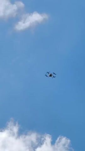 A drone flies in a clear blue sky with scattered white clouds. The drone is small and appears to be a quadcopter with four propellers. It is viewed from below, looking up.