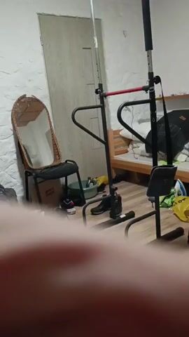 The video opens with a static shot of a room. A pull-up bar stands in the center of the frame, with a wooden chair and a wicker-backed chair to its left. Behind the pull-up bar is a door, and to the right of the pull-up bar is a bed with pillows and blankets. The walls are white and textured, and the floor is made of light-colored wood. In the foreground, a blurred arm enters the frame. The camera then pans slightly to the right, showing more of the bed and some yellow bags. The arm remains in the foreground, obscuring part of the view. The video continues to pan slowly to the right, revealing a black chair and a backpack leaning against the wall. The arm in the foreground continues to block a significant portion of the view. The camera then pans back to the left, showing the wicker chair and the pull-up bar. The arm remains in the foreground. The video continues with the camera slowly panning left and right, the arm in the foreground always present and obscuring the view. There are no discernible actions or interactions taking place other than the slow movement of the camera. The environment appears to be a bedroom or a multi-purpose room with exercise equipment.
