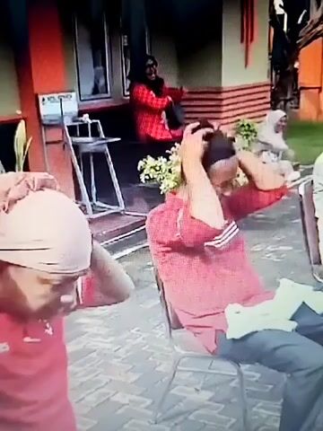 The video shows a group of people sitting on chairs outdoors, possibly in a yard or courtyard. In the foreground, a person wearing a red shirt and a light-colored head covering is bending over, touching their head. To their right, a person in a red shirt and dark pants is seated, holding flowers and touching their head. In the background, several other people are seated, some wearing head coverings. The environment appears to be decorated with red and white flags or banners. The overall scene suggests some sort of gathering or ceremony.