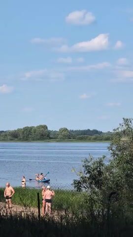 A wide shot shows a serene lake under a clear blue sky with scattered white clouds. Several people are visible in and around the water. In the foreground, three individuals are standing on a sandy area, two in bathing suits. Further back, a person wades in the shallow water. In the middle ground, a blue kayak with at least three people inside is being paddled across the lake. The far bank of the lake is lined with lush green trees. A fence runs across the foreground, partially obscuring the view. The overall impression is a peaceful summer day at a lake.