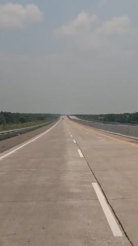 The video shows a long, empty highway with a few cars visible in the distance. The road is concrete and well-maintained, with clear lane markings. There are trees and vegetation on either side of the road, and the sky is partly cloudy. The camera appears to be mounted on a moving vehicle, as the perspective is constantly shifting slightly.