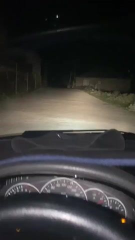 The video shows a first-person perspective from inside a car at night. The dashboard and steering wheel are visible in the foreground. The car is driving down a dimly lit road with houses visible on either side. The headlights of the car illuminate the road ahead. A tablet is mounted on the dashboard, displaying a music app with song information. The scene suggests a drive through a rural or suburban area at night.