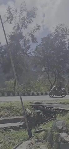 A motorcycle is visible on the left side of the frame, moving along a road that curves to the right. The road has a white and black striped barrier along its edge. Beyond the road, a hillside is covered in trees and vegetation. There is a white object on the hillside, and some smoke or mist is rising from the hillside, obscuring the view of the terrain further up. In the foreground, there is a wooden structure crossing a small ditch or stream. The sky is partly cloudy.