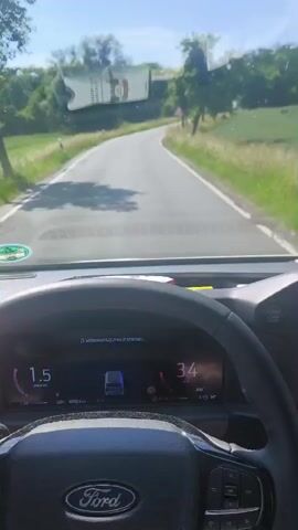 The video shows the interior of a Ford vehicle driving on a rural road. The driver's perspective is captured from the front seats, focusing on the dashboard and the road ahead. On the dashboard, a digital display shows the car's speed, which fluctuates between 35 km/h and 47 km/h. A navigation system on the central screen displays a map with a route plotted out. A smartphone, mounted on a stand, also shows a navigation app with a similar map. The car is surrounded by lush green fields and trees, indicating a sunny day in a countryside setting. The road appears to be paved and curves gently. Traffic signs are visible along the roadside, including a warning sign for a curve ahead. The overall scene depicts a calm and scenic drive through the countryside.