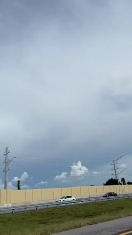 The video shows a highway scene with a cloudy sky. There are several cars and trucks on the road. A fire truck with the words SARASOTA COUNTY FIRE DEPARTMENT ADVANCED LIFE SUPPORT is parked on the side of the road. The fire truck is red and has flashing lights. There is also a white SUV parked behind the fire truck. The fire truck has a ladder on top. There is a concrete barrier along the side of the highway. There are trees and power lines in the background. The scene appears to be a roadside incident.