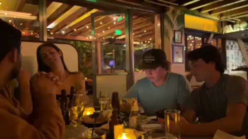 The scene shows four people sitting around a table in what appears to be an outdoor restaurant or patio area. There's a man in a brown jacket facing the camera. To his right is a woman with long, dark hair. Next to her are two men. The man in the middle is wearing a green t-shirt and a baseball cap and looking at a mobile phone. The man on the right is wearing a gray t-shirt. The table is set with glasses, bottles of beer, plates, candles and a basket that looks like it may contain tortilla chips. The setting is decorated with string lights and has tropical-themed artwork. Other patrons can be seen in the background. The overall atmosphere appears relaxed and social.