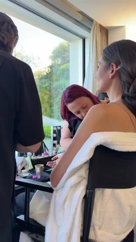 Several people are in a brightly lit room, likely a dressing room, getting ready.  Most of them are seated and wearing white robes. Several makeup artists are working on models, applying makeup or doing manicures. There's a makeup station with various products and tools. Some people are using their phones. Large windows let in natural light, and trees are visible outside. The room seems busy and organized. One person has short purple hair.