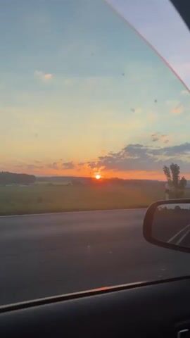The video shows a view from inside a car, looking out the side window. The car is traveling on a road. Outside the window, there is a sunset with an orange and yellow sky and some clouds. There are green fields and some trees along the roadside. The car's side mirror is visible in the lower right corner of the frame, reflecting the road behind. The sun is low on the horizon, casting long rays of light.