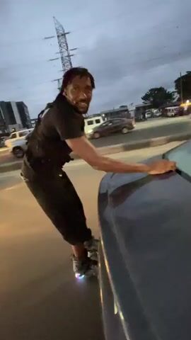 A man on roller skates, wearing a black t-shirt and black pants, leans against the side of a dark-colored car. He is holding onto the car with his right hand. His roller skates have lights on them that are illuminated. In the background, there are cars on a road, streetlights, and power lines. The sky is cloudy and appears to be dusk. Through the rear window of the car, two children are visible. The child on the right is smiling and waving at the camera. The child on the left is also visible and appears to be looking towards the camera. The overall environment suggests an outdoor setting at twilight, possibly a parking lot or a street.