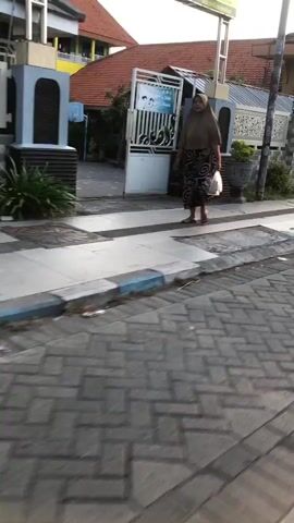 A full shot video shows a woman standing outside a building entrance. The woman is wearing a headscarf and a patterned dress. She is holding a clear plastic bag in her right hand. The entrance appears to be to a school, with a sign above the door. The building has red tiled roofs. The ground is paved with bricks. The lighting suggests it is daytime, possibly late afternoon, due to the angle of the shadows.