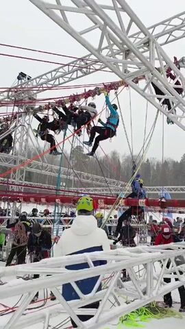 The video shows a group of people participating in a ropes course in a snowy outdoor environment. The course is a complex structure of metal frames and ropes, with participants wearing harnesses and helmets. Some participants are actively navigating the course, suspended in the air and moving along the ropes. Many people are standing below, presumably waiting their turn or observing. The ground is covered in snow, and bare trees are visible in the background. The weather appears overcast.
