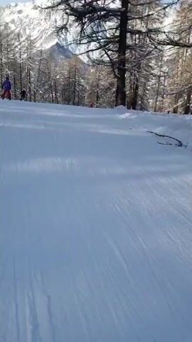 The video shows a first-person perspective of someone snowboarding down a snowy slope. Several other skiers and snowboarders are visible on the slope, and the scenery includes snow-covered trees and a large, snow-capped mountain in the background under a partly cloudy sky. The snowboarder's board is visible at times and has the word "Sila" written on it.