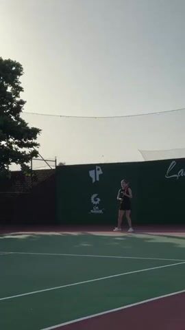 A woman in athletic wear is playing tennis on an outdoor court. She is positioned to hit a forehand shot with a tennis racket. The court has green and red surfaces, with white lines. A green wall with logos is in the background. The sky is bright and hazy. A tennis ball is visible in the air.