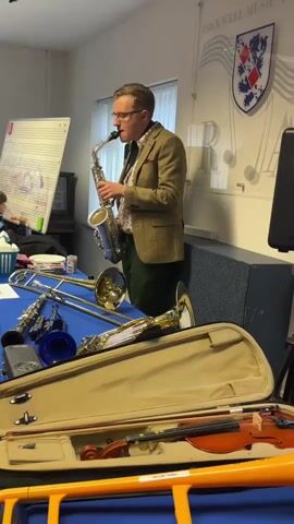 A man in a tweed jacket and glasses plays a saxophone in a room with musical instruments on a blue table. A whiteboard with musical notation is visible on the left. A violin case is in the foreground. The man is standing, facing right, and blowing into the saxophone. Several brass instruments, including a trombone, are laid out on the table. There is a logo on the wall behind the man that appears to be a crest with musical notes. The room is well-lit.