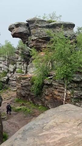 The video shows a person walking on a muddy path between large rock formations. The rocks are layered and have vegetation growing on them. Trees, including birch trees, are visible in the background. The sky is overcast, suggesting a cloudy or rainy day. The video pans across the rocky landscape, highlighting the scale and texture of the rock formations and the surrounding forest.