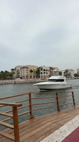 A white yacht with a black hull stripe and a radar dome on its roof moves to the right across a body of water. It is passing a pier made of light brown wood with wooden railings in the foreground. In the background, behind the yacht, are buildings with light-colored facades, arched windows, and red-tiled roofs, with palm trees visible between them. The sky is overcast and gray.