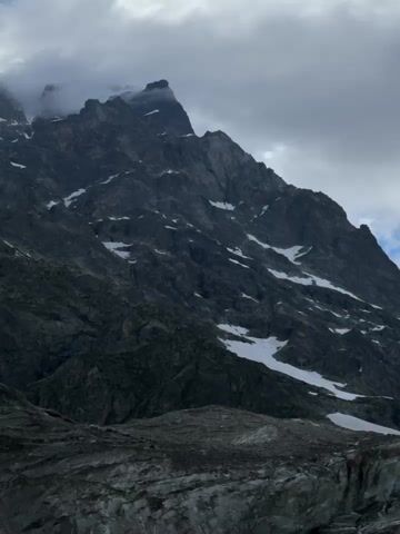 A video shows a rocky mountain landscape with snow patches and a glacier in the foreground. A stream flows down the rocks. The sky is blue with scattered clouds. The camera pans across the scene.