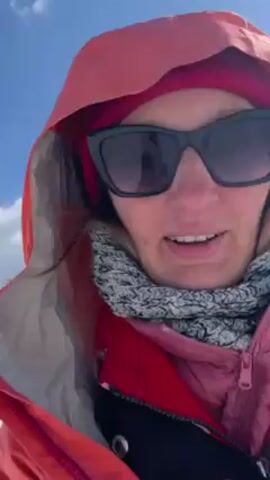 The video shows a person wearing sunglasses and a red jacket, standing in a snow-covered environment with a blue sky and some clouds. The person yells "Woohoo!", and then skis downhill. The snow covered hills, trees and mountains can be seen. The person casts a shadow on the snow.