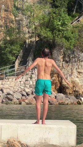A man with a bare torso, wearing bright green swim trunks, stands facing away from the camera on a concrete pier. He is moving his arms in front of him, possibly stretching. Behind him is a rocky cliff with trees and a stone staircase with a metal railing leading up the cliffside. The water is calm and slightly reflective. The scene is bathed in bright sunlight.