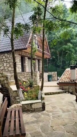 The video shows a stone house nestled in a lush, green landscape, with flowers blooming in colorful pots and a wooden bench in the foreground. Smoke rises from a chimney, hinting at a warm, inviting atmosphere. The camera slowly pans left, revealing more of the surrounding nature and another stone house in the background.