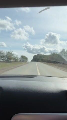 A vehicle travels on a two-lane road on a partly cloudy day. The road has a yellow center line. Trees line both sides of the road. The dashboard of the vehicle is visible in the foreground. The sky is visible through the windshield.