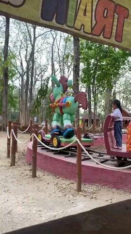A small child rides on a toy train attraction in an outdoor park. The train car the child is in is a red train car with wooden accents and a large pink wheel. The child, wearing a white shirt and blue pants, is facing away from the camera. In front of the child's car is a green, cartoonish horse figure with a red saddle and blue boots, also on its own train car. The attraction appears to be a miniature train track in a dirt area surrounded by trees and a rope barrier. A yellow banner with colorful letters, partially visible and appearing to say "TRAIN," hangs above the attraction. The video captures the train moving forward.